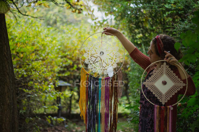 Femme en tenue décontractée montrant des attrape-rêves faits à la main avec de longs fils regardant l'amulette passer du temps en plein air — Photo de stock