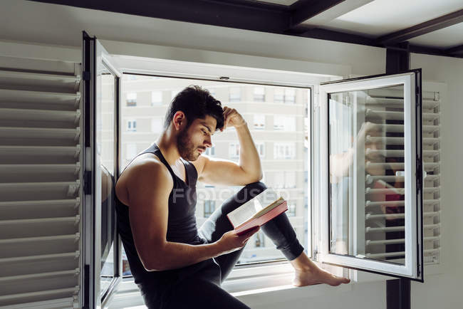 Vista laterale di giovane uomo bello serio in camicia casual senza maniche seduto sulla finestra e la lettura di libro interessante — Foto stock
