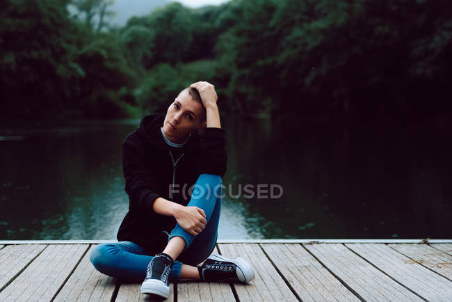 Female hipster with short hairstyle in casual clothing sitting with crossed legs on wooden pier of pond with green trees on blurred background — Stock Photo