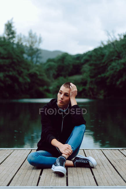 Hipster femenino con peinado corto en ropa casual sentado con las piernas cruzadas en el muelle de madera de estanque con árboles verdes sobre fondo borroso - foto de stock