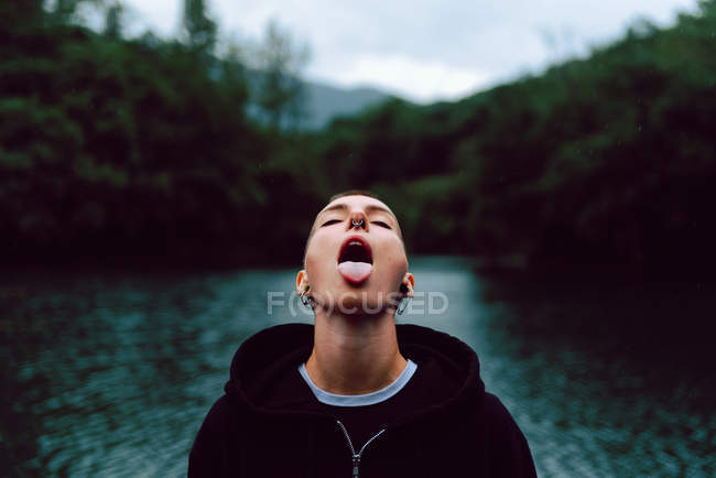 Short haired woman with piercing wearing black hoodie looking up while catching raindrops with tongue near green forest and pond — Stock Photo