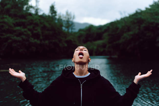 Short haired woman with piercing wearing black hoodie and spreading hands while catching raindrops with tongue near green forest and pond — Stock Photo