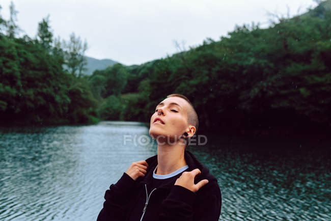 Frau mit Kurzhaarfrisur und Piercing in lässiger Kleidung mit geschlossenen Augen zum Himmel mit Teich inmitten grüner Pflanzen auf verschwommenem Hintergrund — Stockfoto