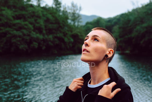 Femme avec coiffure courte et perçage dans des vêtements décontractés regardant vers le ciel avec étang parmi les plantes vertes sur fond flou — Photo de stock