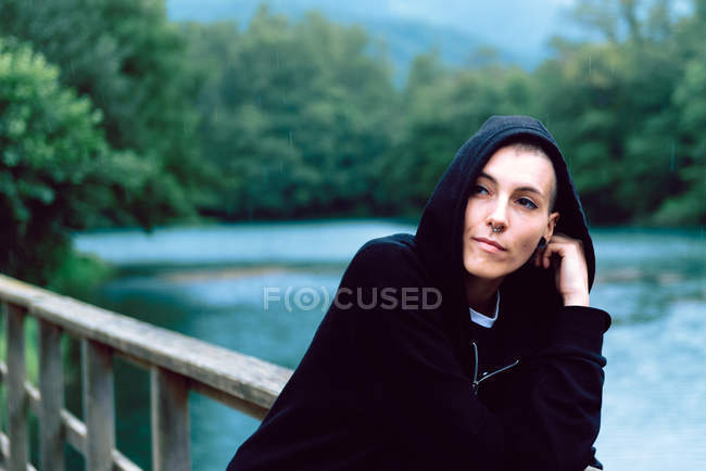 Woman in black hoodie leaning on wooden fence of bridge and looking away with turquoise pond and green plants on blurred background — Stock Photo