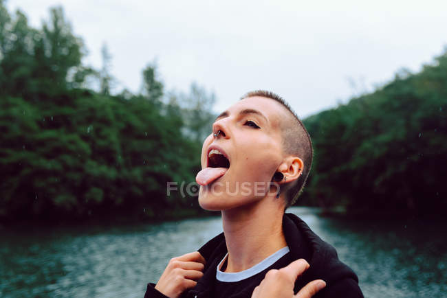 Mulher com piercing vestindo capuz preto olhando para cima enquanto captura gotas de chuva com língua perto de floresta verde e lagoa — Fotografia de Stock