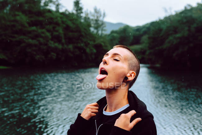Woman with piercing wearing black hoodie looking up while catching raindrops with tongue near green forest and pond — Stock Photo