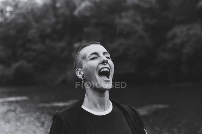 Frau mit Piercing trägt schwarzen Kapuzenpulli und schaut auf, während sie Regentropfen mit Zunge in der Nähe von grünem Wald und Teich fängt — Stockfoto