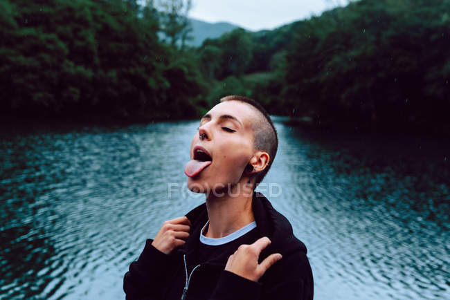 Жінка з пірсингом в чорному светрі, дивлячись вгору, ловить дощові пагони з язиком біля зеленого лісу і ставка — стокове фото
