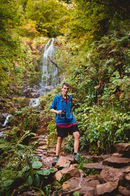 Жінка з професійною камерою і рюкзаком, дивлячись далеко, стоячи на водоспаді в лісі в літній день — стокове фото