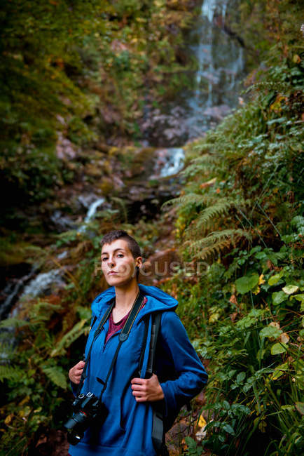 Зосереджена молода жінка з професійною камерою і рюкзаком, дивлячись на камеру, стоячи на водоспаді в лісі в літній день — стокове фото