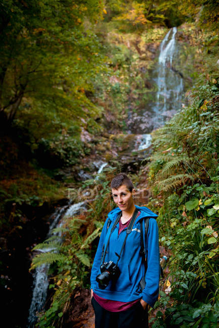 Mujer joven enfocada con cámara profesional y mochila mirando a la cámara mientras está de pie en la cascada en el bosque en el día de verano - foto de stock