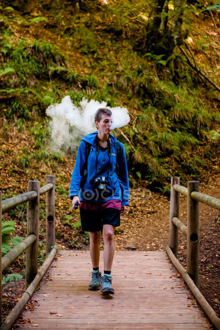 Femme avec caméra debout sur un pont en bois derrière une épaisse fumée tout en fumant émoussé pendant le camping en forêt — Photo de stock