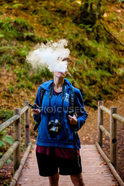 Faceless woman with camera standing on wooden bridge behind thick smoke while smoking blunt during camping in forest — Stock Photo
