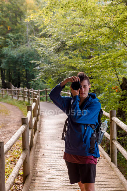 Caminhante sério desfrutando de férias e tirando foto na câmera profissional enquanto está em pé no caminho de madeira na floresta — Fotografia de Stock