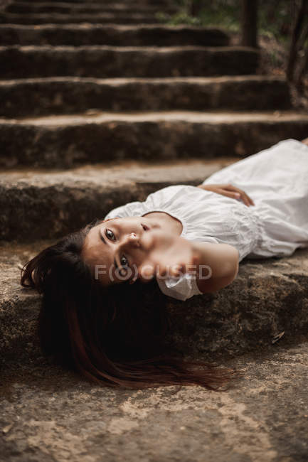 Desde arriba de tierna mujer joven encantadora en vestido blanco acostado en las escaleras en el parque otoñal mirando a la cámara alcanzando con la mano - foto de stock