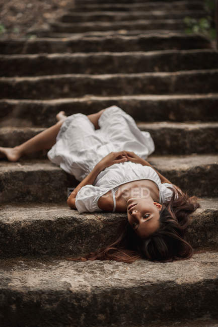 D'en haut de tendre charmante jeune femme en robe blanche couchée vulnérablement sur les escaliers dans le parc automnal regardant loin — Photo de stock