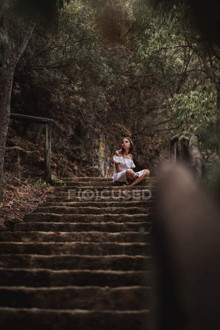 From above of tender charming young woman in white dress sitting on stairs in autumnal park looking away — Stock Photo