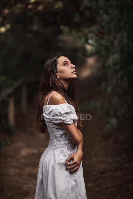 Vista laterale di tenera splendida giovane donna in abito bianco che abbraccia le spalle in piedi in un parco calmo e guardando altrove — Foto stock