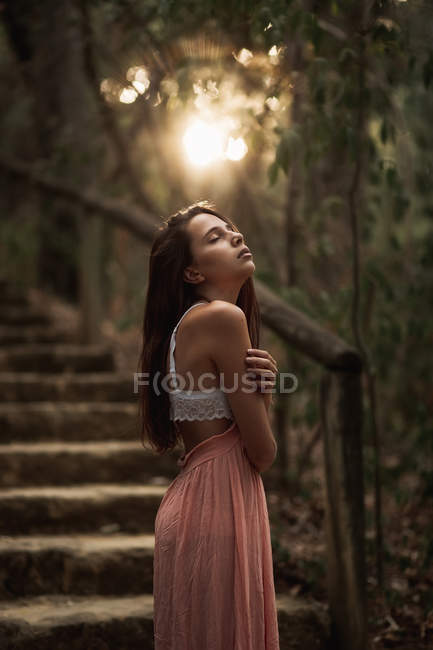 Vista lateral da mulher suave em vestido rosa e sutiã de renda branca em pé no parque outonal com os olhos fechados em volta iluminado — Fotografia de Stock