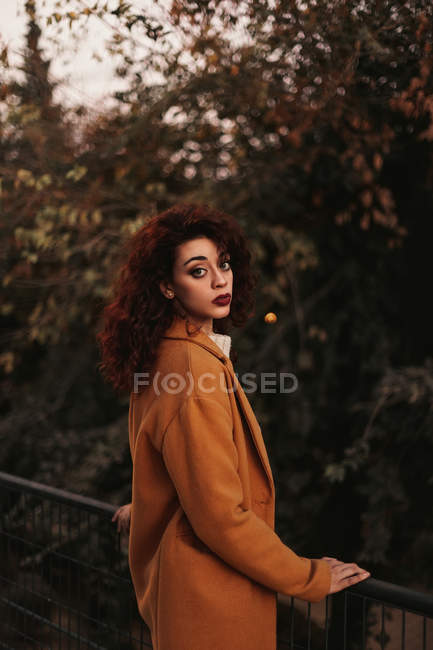 Женщина с темными кудрявыми волосами в трикотажном свитере и пальто, стоящие в парке, кладущие руку на металлические перила, глядя в камеру — стоковое фото