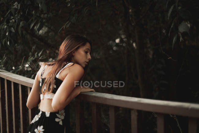 Vista posteriore della donna tenera in abito elegante appoggiato sulla recinzione di legno e guardando verso il basso in un parco tranquillo — Foto stock