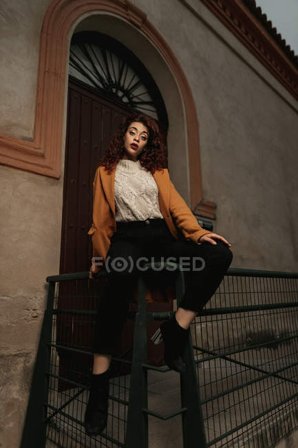 Woman in overcoat and high heeled boots sitting on metal fence next to building door while looking at camera with challenge — Stock Photo