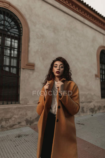 Donna in maglia calda maglione e cappotto con le braccia alzate in piedi accanto all'edificio mentre guarda la fotocamera con sfida — Foto stock
