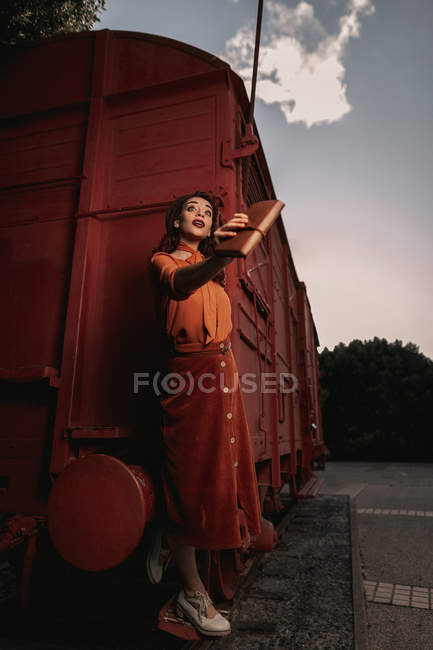 Femme aux cheveux bouclés foncés en béret portant des vêtements en terre cuite de style vintage dans le dos éclairé debout sur l'étape du train de voiture — Photo de stock