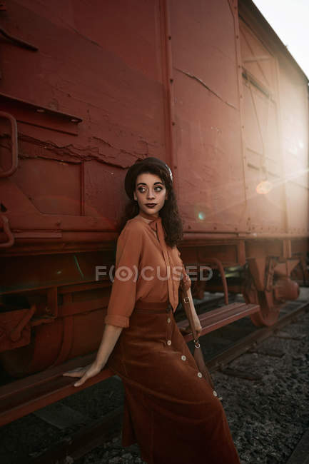 Woman with dark curly hair in beret wearing terracotta clothes in vintage style in back lit sitting on step of car train — Stock Photo