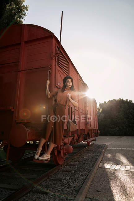 Femme aux cheveux bouclés foncés en béret portant des vêtements en terre cuite de style vintage dans le dos éclairé debout sur l'étape du train de voiture — Photo de stock