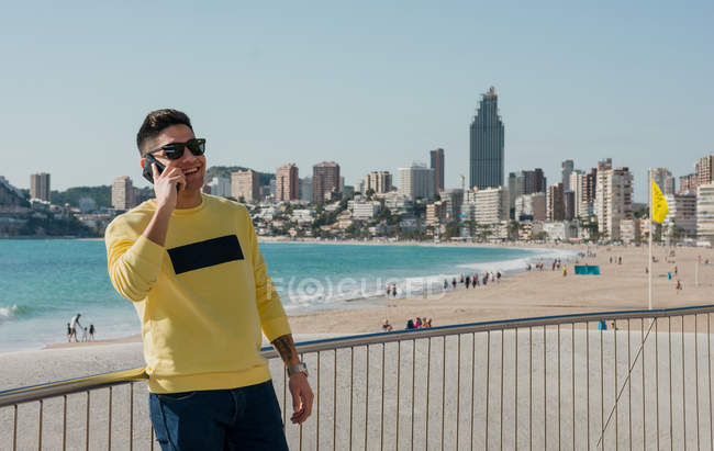Людина в повсякденному одязі і модні сонцезахисні окуляри розмовляють на мобільному телефоні на набережній з сучасним міським пейзажем на фоні — стокове фото