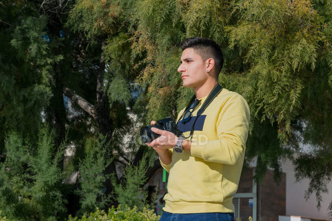 Низкий угол задумчивого мужчины-фотографа с черными волосами в повседневной одежде, стоящего с фотокамерой и смотрящего в зеленый парк — стоковое фото