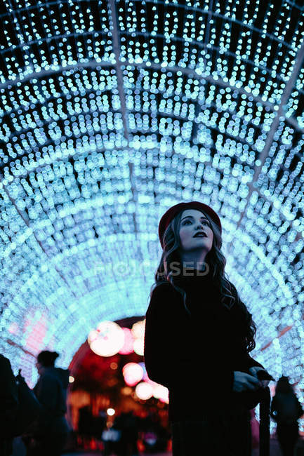 Elegant woman in trendy clothing and hat near wall with lights in Christmas time — Stock Photo