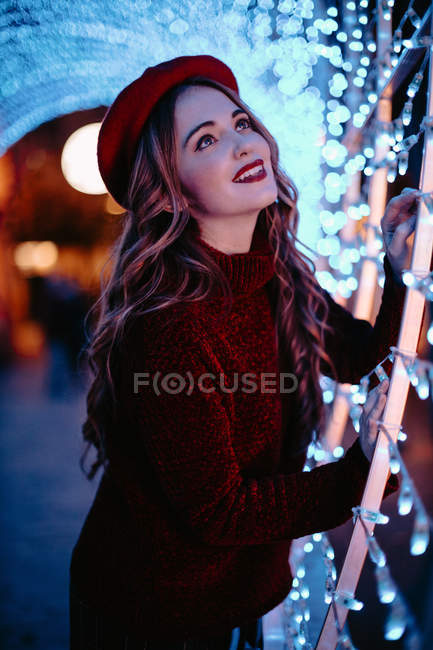 Элегантная женщина в модной одежде и шляпе возле стены со светом в Рождество — стоковое фото
