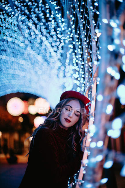 Элегантная женщина в модной одежде и шляпе возле стены со светом в Рождество — стоковое фото