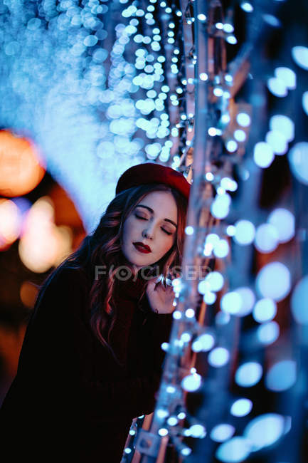Femme élégante en vêtements à la mode et chapeau près du mur avec des lumières pendant la période de Noël — Photo de stock