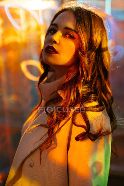Sensual young woman in trendy coat standing in light of neon signs at city street — Stock Photo