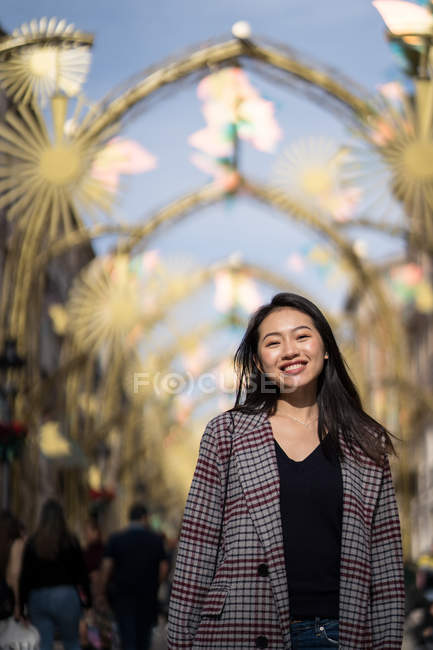 Низкий угол азиатского туриста в черной рубашке и клетчатой куртке, улыбающегося в камеру, стоя на улице против золотых арк — стоковое фото