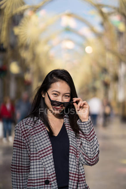 Affascinante donna asiatica in abiti casual e occhiali da sole sorridente alla fotocamera mentre in piedi sulla strada contro archi dorati — Foto stock