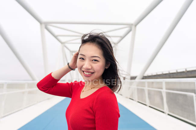 Encantadora mujer asiática con camisa roja sonriendo y mirando a la cámara mientras camina por el moderno puente de la ciudad - foto de stock