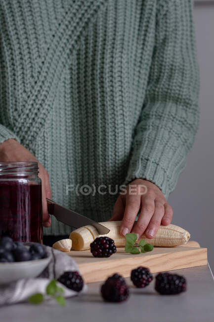 Corte com faca banana fresca em tábua de corte de madeira enquanto prepara o batido de vitamina saudável com amora e mirtilo em casa — Fotografia de Stock