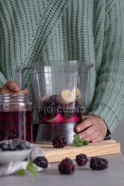 Crop person preparing healthy vitamin smoothie with banana and other berries in combination with mint using blender standing on wooden cutting board on table in modern kitchen — Stock Photo