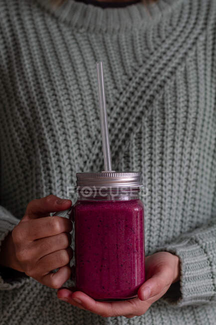 Crop person in casual knitted sweater holding transparent jar with metal lid and plastic straw of tasty vitamin pink beverage of sweet fresh fruits and berries while having healthy appetite breakfast — Stock Photo