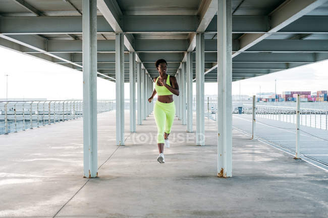 Desde abajo, la deportista afroamericana adulta en vibrante ropa deportiva verde se centra y corre sola a lo largo del paseo marítimo entre columnas de metal bajo el techo. - foto de stock