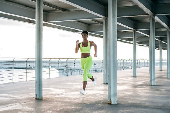 Femme de sport adulte afro-américaine en vêtements de sport vert vif se concentrant et courant seul le long du front de mer parmi les colonnes métalliques sous le toit — Photo de stock