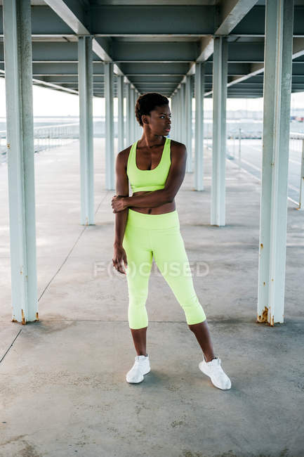 Afroamericana deportista adulta en vibrante ropa deportiva verde enfocándose en pie solo a lo largo del paseo marítimo entre columnas de metal bajo techo - foto de stock