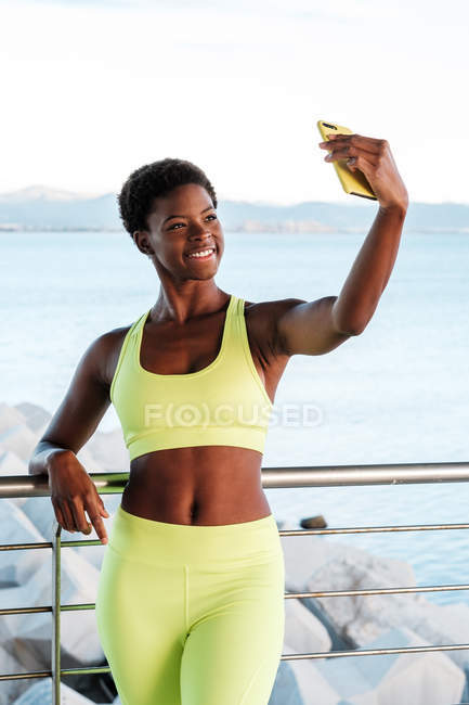 Afrikanische Amerikanerin in leuchtend gelber Sportbekleidung, die sich auf den Bildschirm konzentriert und ein Selfie mit dem Smartphone macht, während sie auf einem Metallzaun an der Promenade vor verschwommener Meereslandschaft steht — Stockfoto