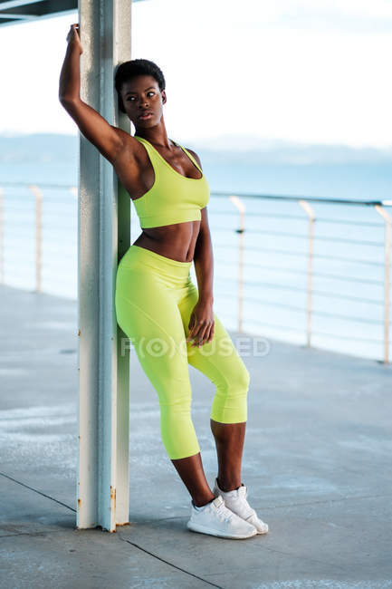 Alluring African American adult woman in bright yellow sportswear looking away and contemplating while standing alone leaning on metal pillar on promenade against blurred seascape in daylight — Stock Photo