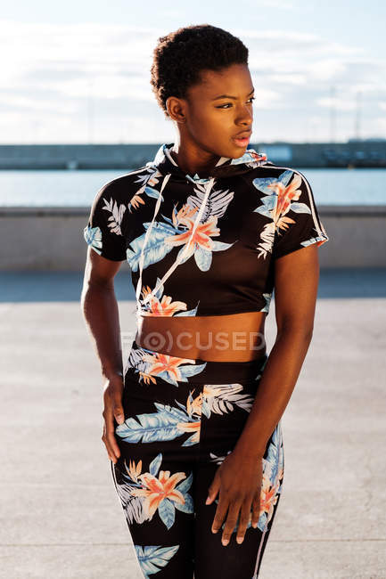 Афроамериканка в цветущем спортивном костюме смотрит в сторону и думает, стоя с руками на бедрах в одиночестве в солнечных лучах против размытой набережной — стоковое фото